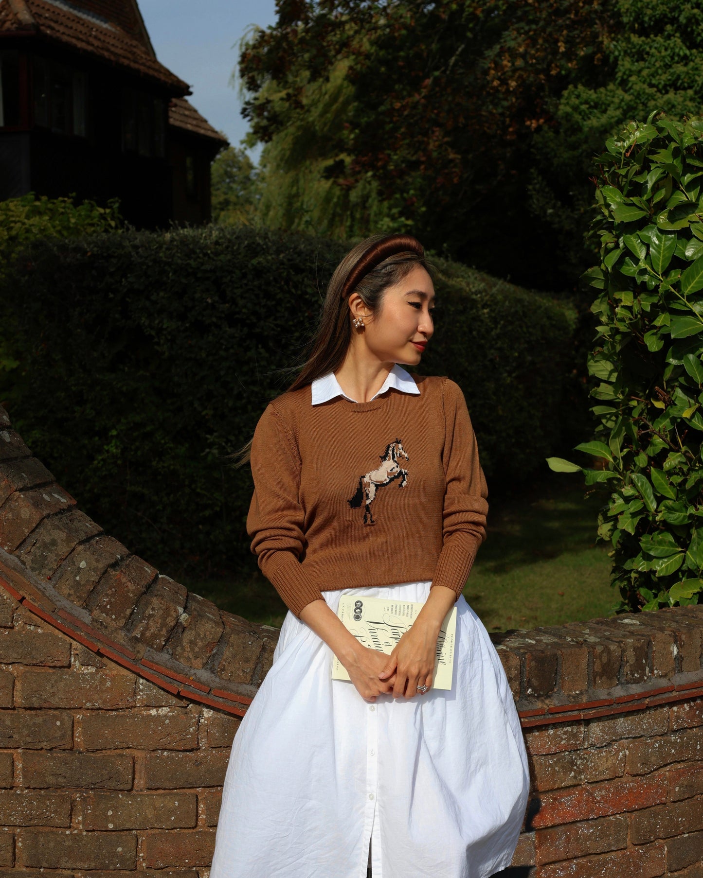 Woman wearing a brown sweater with a horse design, white shirt, and white skirt standing outdoors.