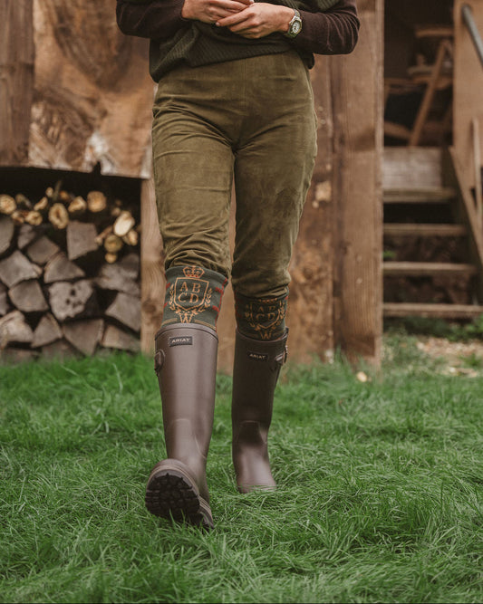 A person walking outdoors on grass in front of a rustic wooden structure, wearing an olive green top, olive pants, and dark brown rain boots with decorative cuffs.