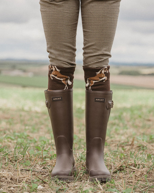 A person standing outdoors in a field wearing brown rain boots with horse illustrations around the cuffs and brown patterned pants.