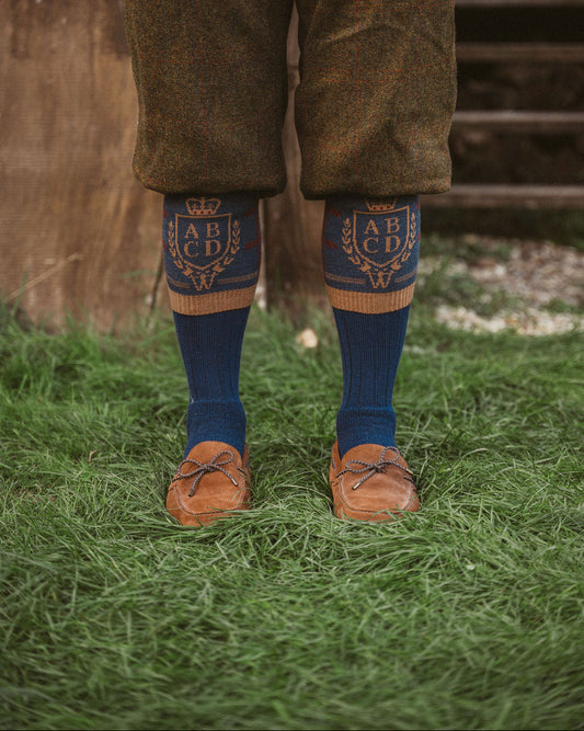 A person standing outdoors on grass wearing brown tweed trousers, dark blue socks with gold embroidered lettering and a crown motif, and brown leather loafers.