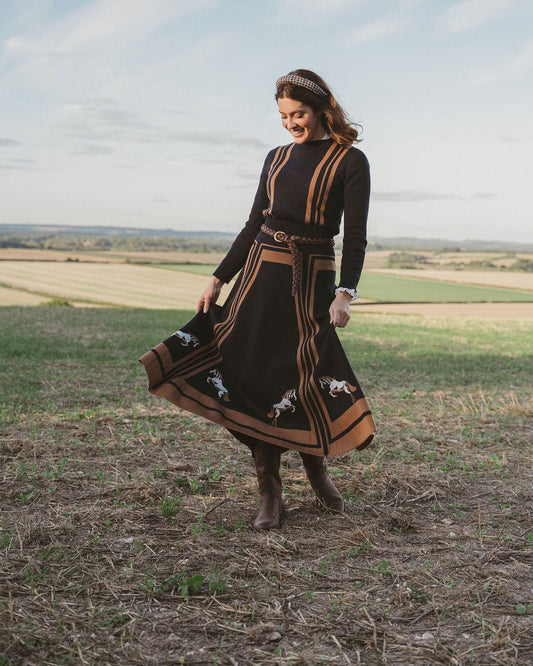 A woman wearing a long-sleeve black midi dress with brown trim and embroidered details, standing in a grassy field under a cloudy sky.