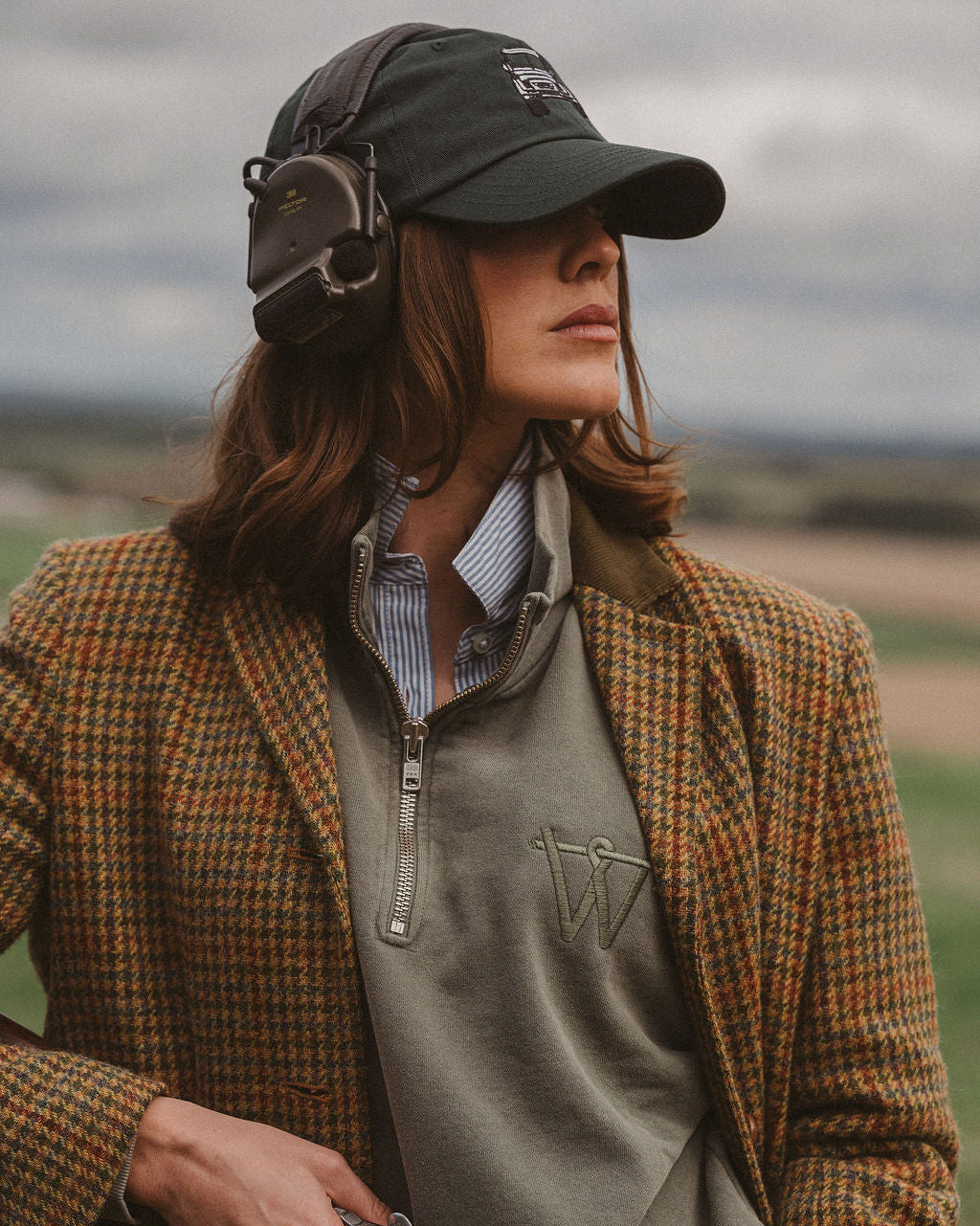 A woman outdoors wearing a brown and orange checkered tweed blazer, a gray quarter-zip sweater, a striped shirt, a dark green cap, and over-ear headphones, holding a shotgun. The background shows a blurred landscape under an overcast sky.