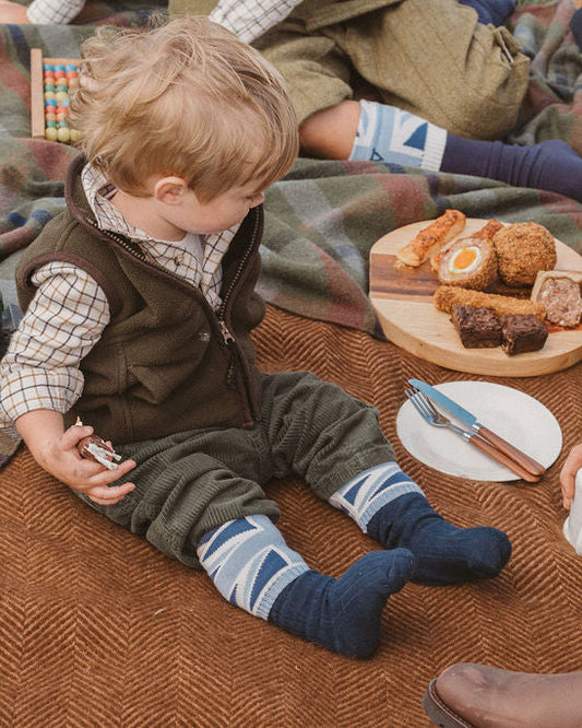A toddler sits on a brown herringbone blanket during a picnic, wearing a brown vest, green trousers, and blue striped socks. A wooden toy tractor, a picnic basket, and a plate with food are nearby.