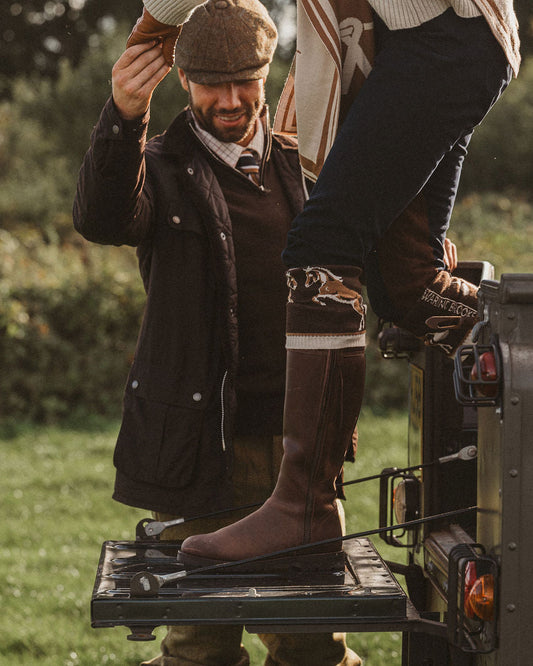 A person wearing tall brown riding boots and a brown flat cap stands on the tailgate of a vehicle outdoors while another person adjusts the hat. The scene is styled in a rustic, outdoor setting with grass and trees in the background.