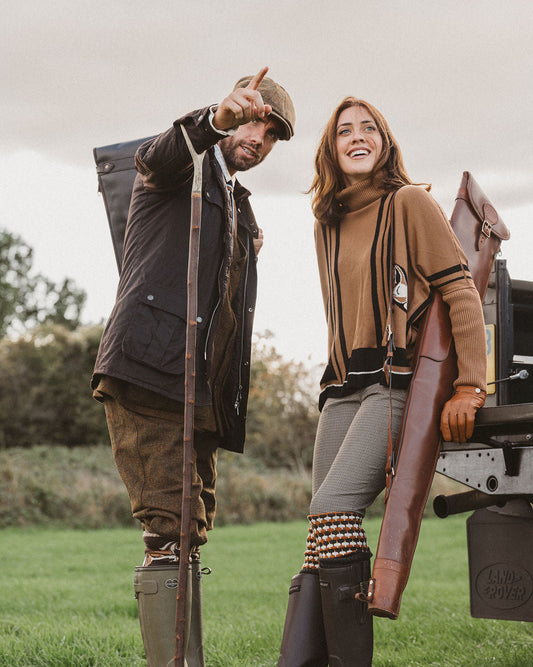 A man and a woman in outdoor hunting attire standing in a grassy field beside a vehicle. The man wears a brown jacket, trousers, tall rubber boots, and holds a shotgun.