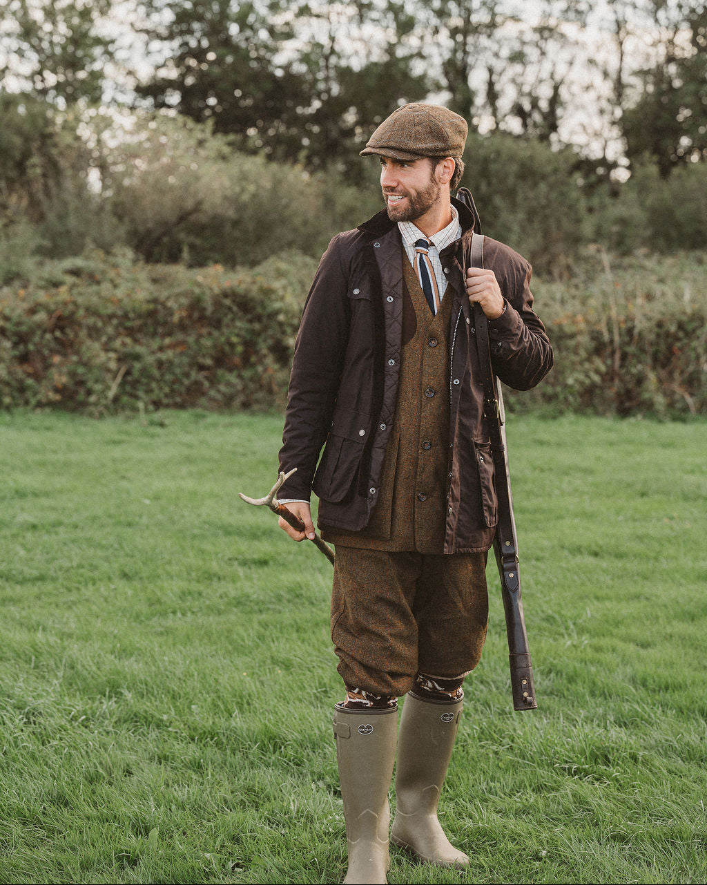 A man wearing a vintage hunting outfit stands in a grassy field. He is dressed in a brown flat cap, dark brown waxed jacket, brown waistcoat, brown corduroy trousers, a patterned shirt, a tie, and tall beige rubber boots, while holding a shotgun.
