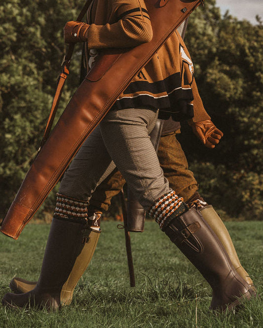 A person walking outdoors on grass wearing tall dark rubber boots, striped knee-high socks, and a brown jacket, while carrying a large brown leather bag or case.