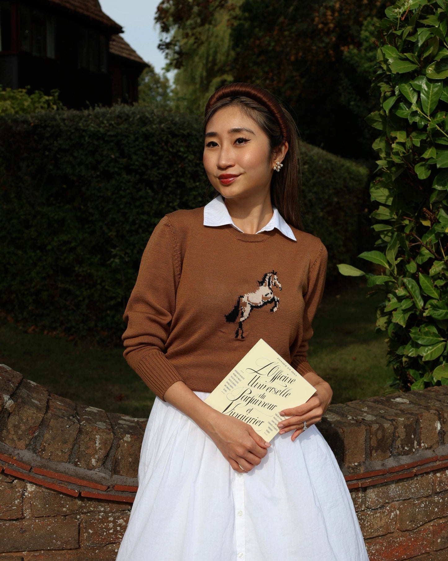 Woman wearing a brown sweater with a horse design, holding a book, standing outdoors.