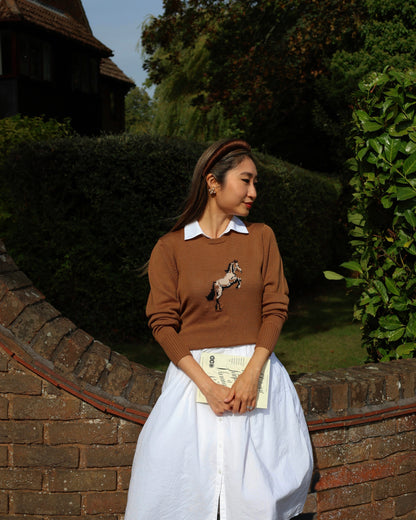 Woman wearing a brown sweater with a horse design, white shirt, and white skirt standing outdoors.