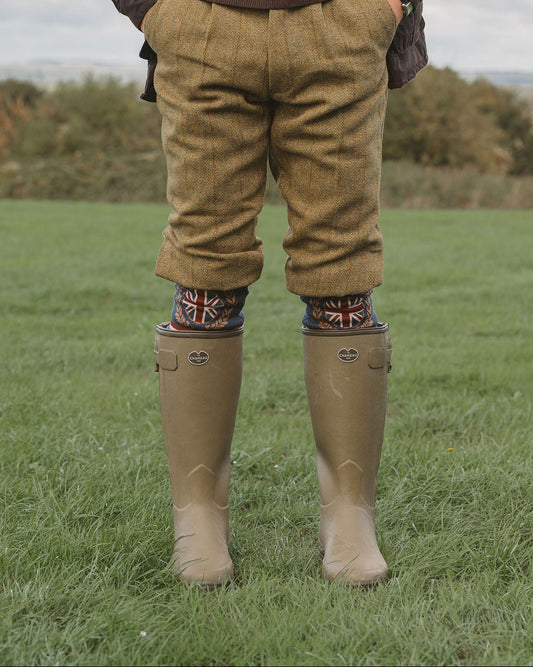 A person standing outdoors on grass wearing brown tweed trousers, a dark brown sweater, a dark jacket, patterned socks, and beige rubber boots.
