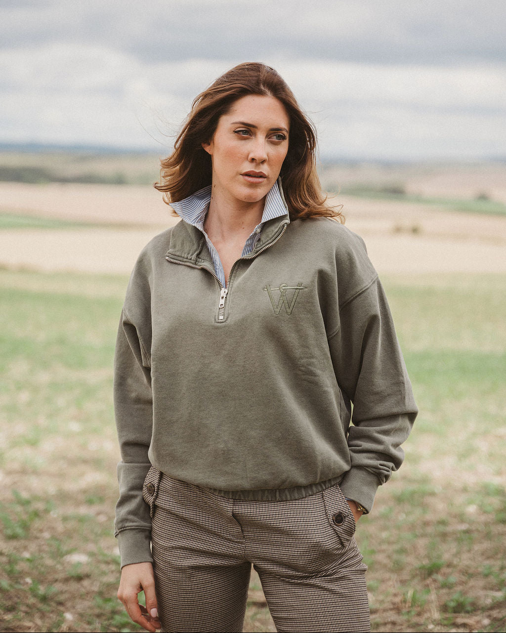 A woman standing in an open field wearing an olive green quarter-zip pullover sweatshirt and matching brown pants under a cloudy sky.