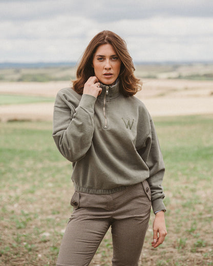 A woman standing in an open field wearing a muted olive-gray quarter-zip pullover sweatshirt with a small embroidered letter 'W' on the chest, paired with matching brown pants.