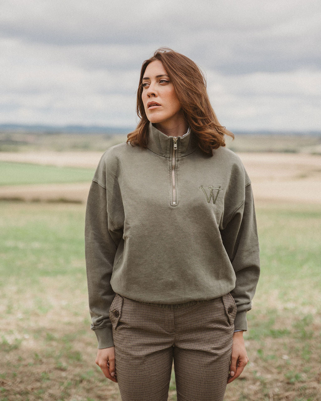 A woman standing outdoors in a field wearing an olive green quarter-zip pullover sweatshirt with a tonal embroidered 'W' logo on the chest, paired with matching olive pants.