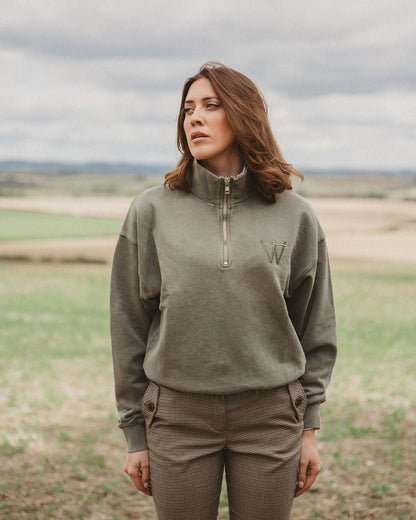 A woman standing outdoors in a field wearing an olive green quarter-zip pullover sweatshirt with a tonal embroidered 'W' logo on the chest, paired with matching olive pants.