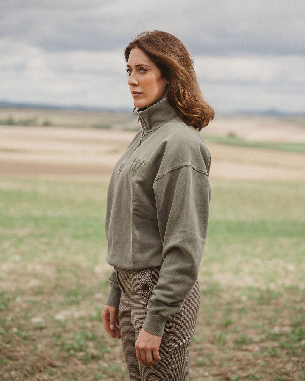 A woman standing in a field wearing an olive green quarter-zip fleece pullover and matching brown pants, photographed outdoors under a cloudy sky.