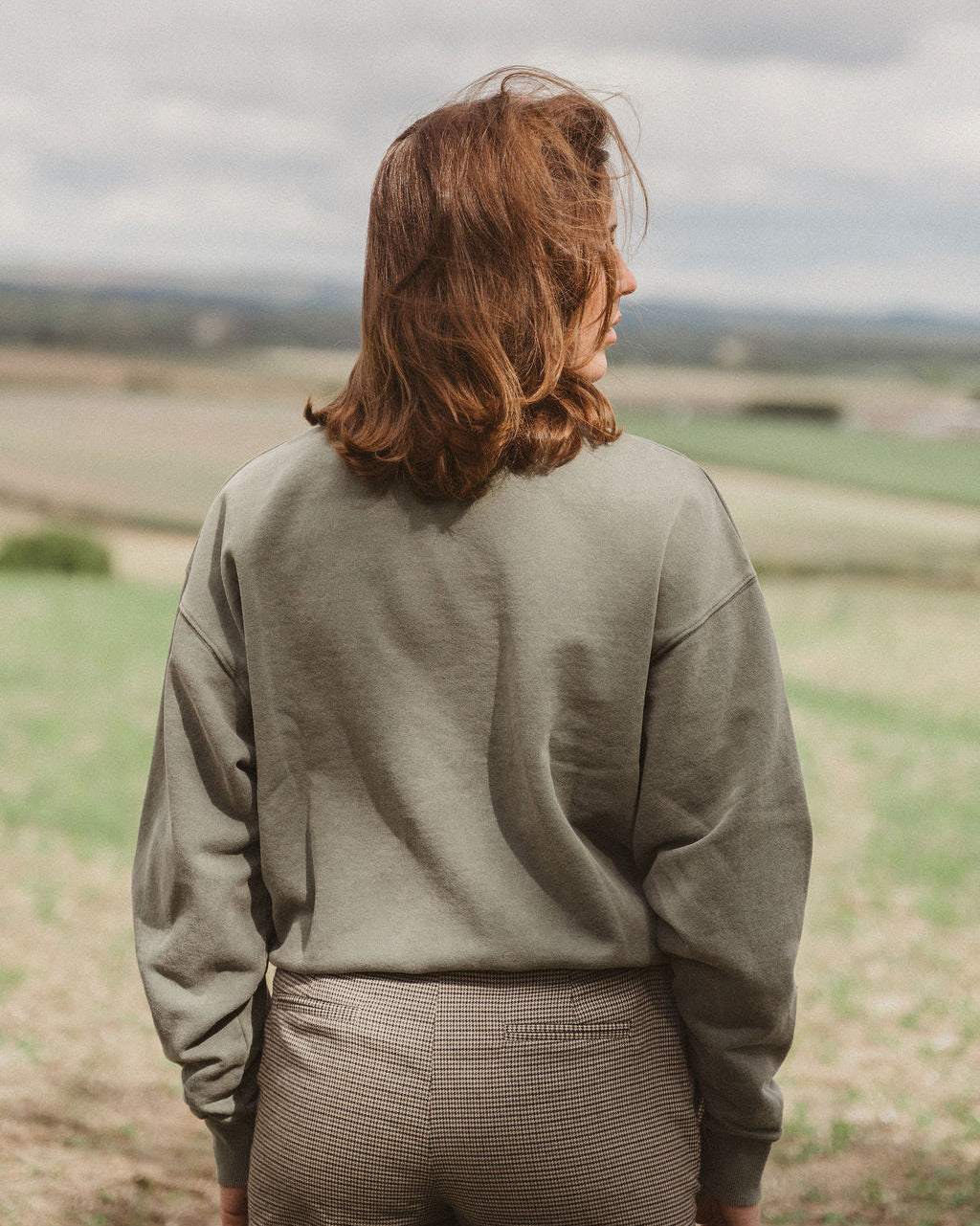 A woman with shoulder-length auburn hair stands outdoors in a field, viewed from behind, wearing an olive green sweatshirt and brown textured pants under a cloudy sky.