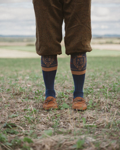 A person standing outdoors in a field wearing brown knee-length corduroy trousers, dark navy socks with gold embroidered monograms, and brown leather shoes.