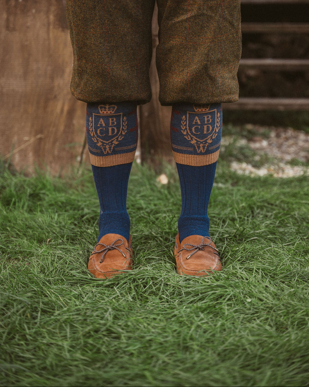 A person standing outdoors on grass wearing brown tweed trousers, dark blue socks with gold embroidered lettering and a crown motif, and brown leather loafers.