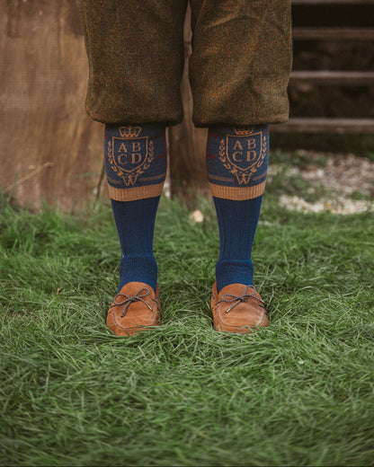 A person standing outdoors on grass wearing brown tweed trousers, dark blue socks with gold embroidered lettering and a crown motif, and brown leather loafers.