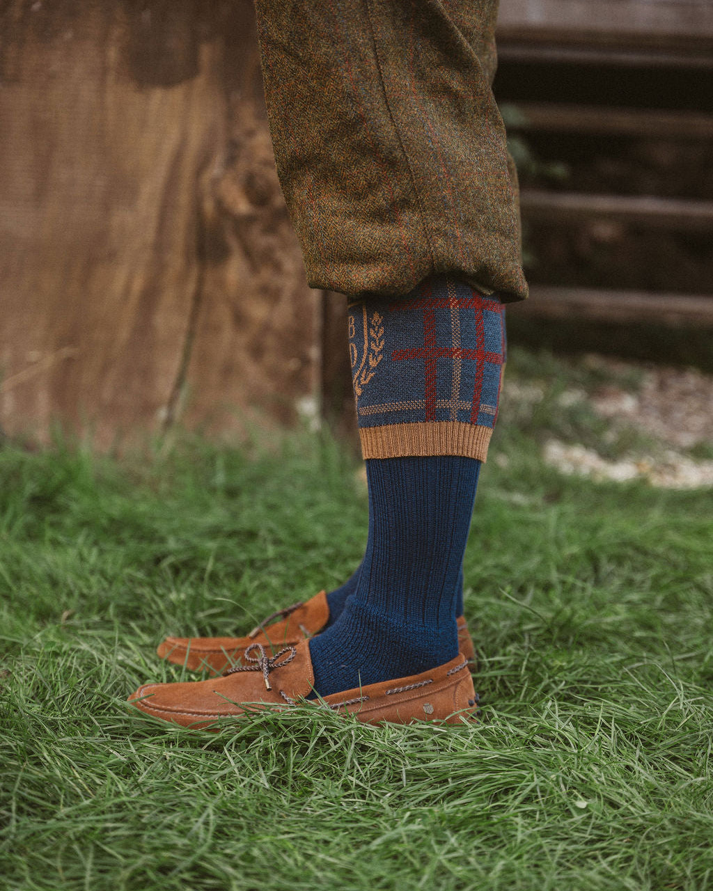 A close-up of a person's lower legs wearing brown tweed trousers, navy blue knit socks with a red and blue plaid pattern and wheat motifs, and brown leather boat shoes, standing on green grass.
