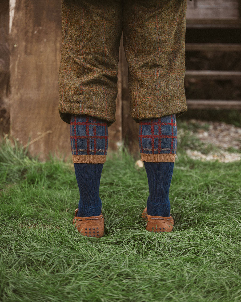 A person standing outdoors on grass wearing brown tweed knee-length trousers, blue socks with a red and blue plaid cuff, and brown leather shoes.