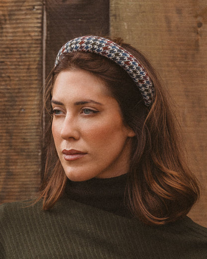 A woman with brown hair wearing a patterned houndstooth headband and a dark olive green turtleneck sweater, posed against a rustic wooden plank background.