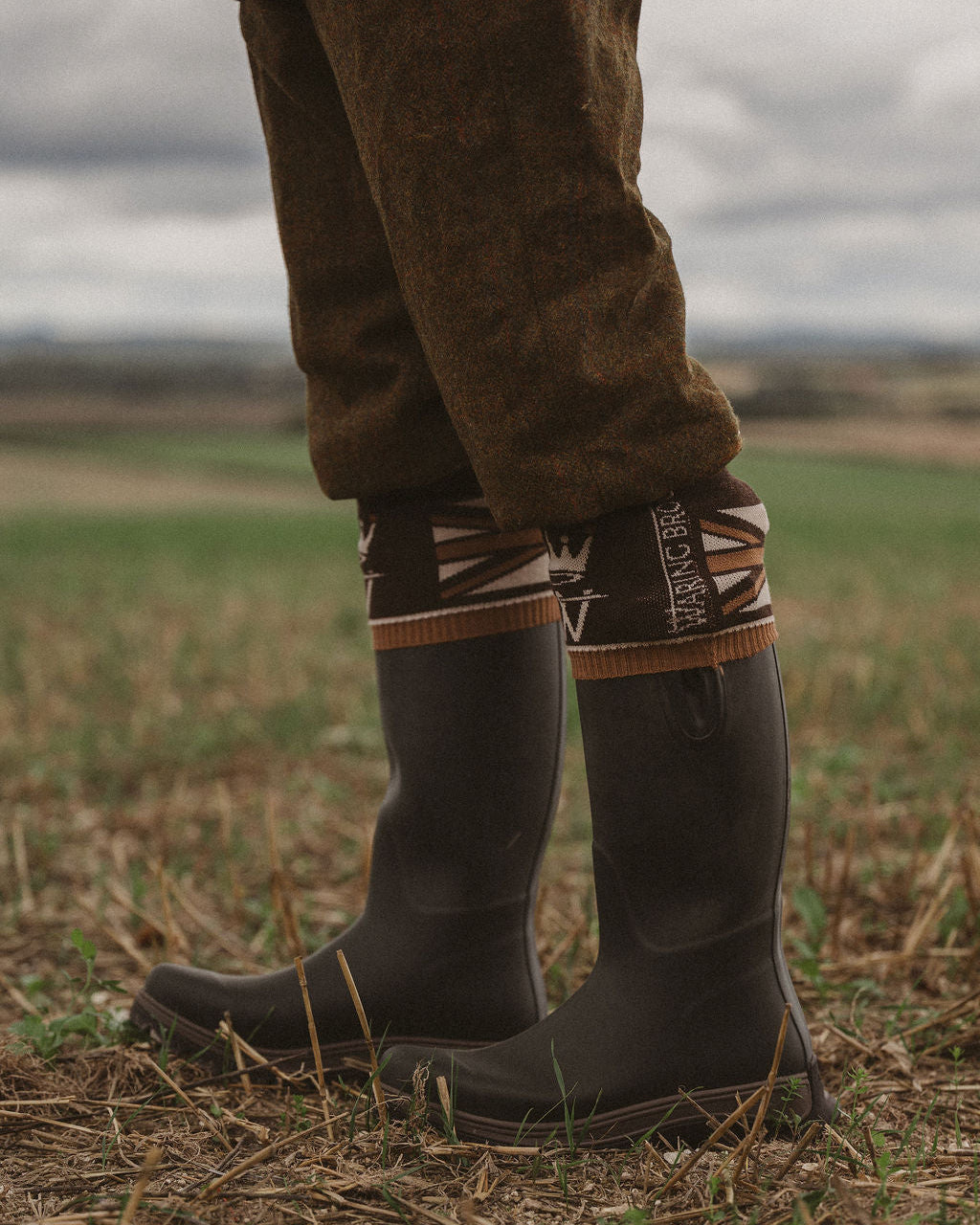 A close-up of a person's legs wearing dark gray rubber rain boots with a patterned brown and beige cuff, standing in a grassy field under an overcast sky.
