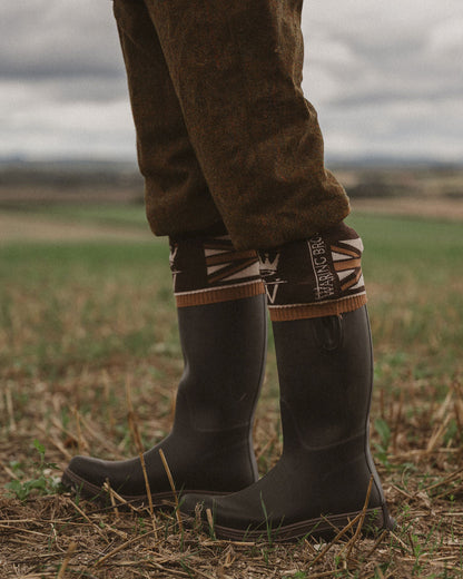 A close-up of a person's legs wearing dark gray rubber rain boots with a patterned brown and beige cuff, standing in a grassy field under an overcast sky.