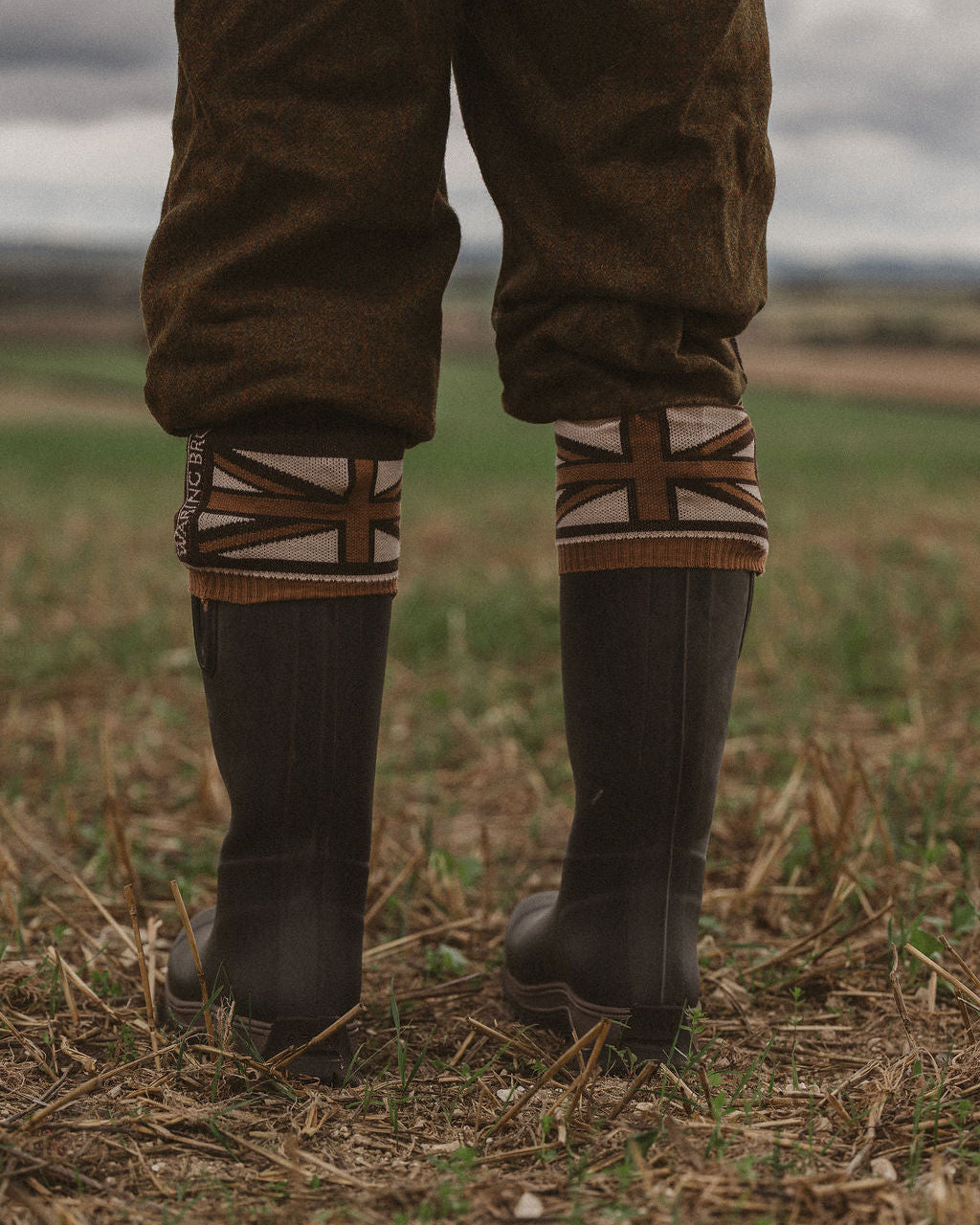 A person standing outdoors in a field wearing dark rubber boots, brown trousers, and knit leg warmers with a Union Jack pattern.