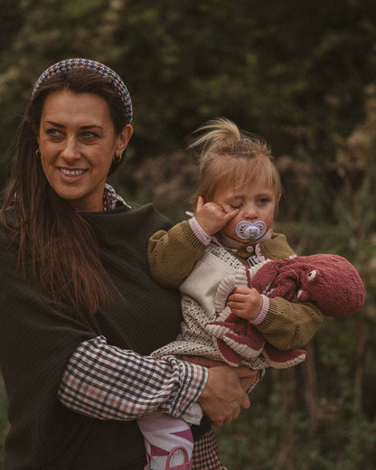 A woman outdoors holding a young child wearing a brown knit sweater, cream vest, and pink booties, with the woman wearing a dark green top and a plaid headband.