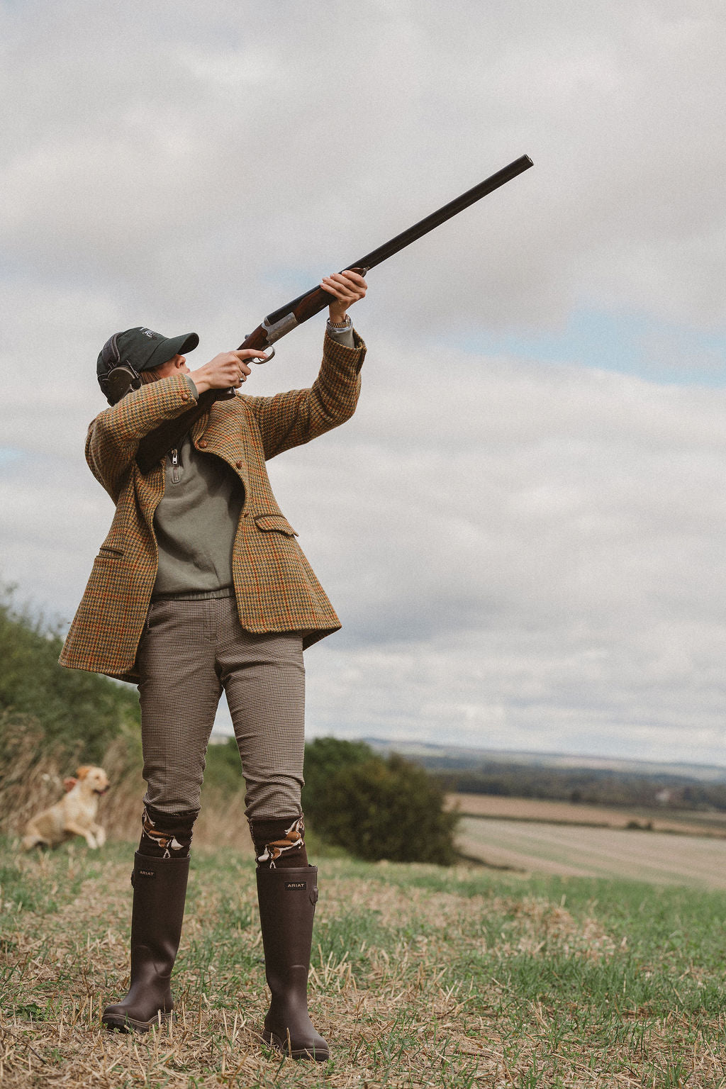 A person in a brown tweed blazer, olive green shirt, brown trousers, and tall brown boots aims a long gun upward in an open field under a cloudy sky. A dog is visible in the background.