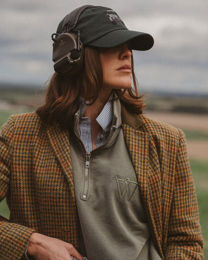 A woman outdoors wearing a brown and orange checkered tweed blazer, a gray quarter-zip sweater, a striped shirt, a dark green cap, and over-ear headphones, holding a shotgun. The background shows a blurred landscape under an overcast sky.