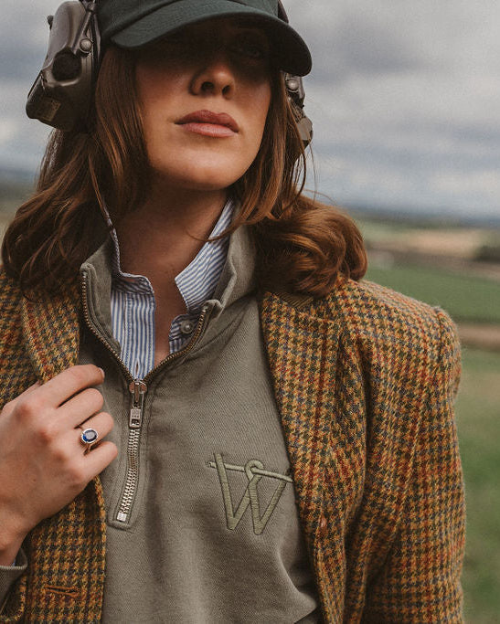 A woman outdoors wearing a brown and tan houndstooth blazer over a taupe quarter-zip pullover, a striped button-down shirt, a dark cap, and over-ear headphones, with a blurred rural landscape in the background.