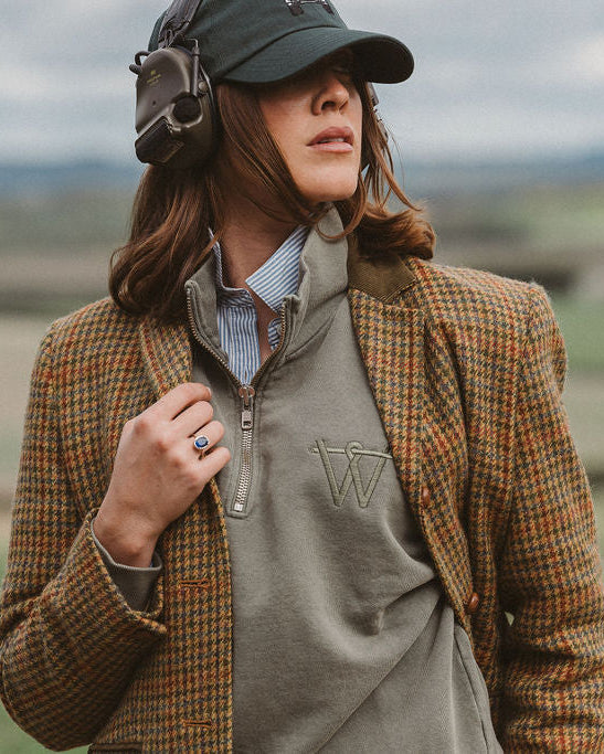A woman outdoors wearing a brown and orange checked tweed blazer over a gray quarter-zip pullover, with a blue striped shirt underneath, a dark cap, and over-ear headphones. The background shows a blurred rural landscape.
