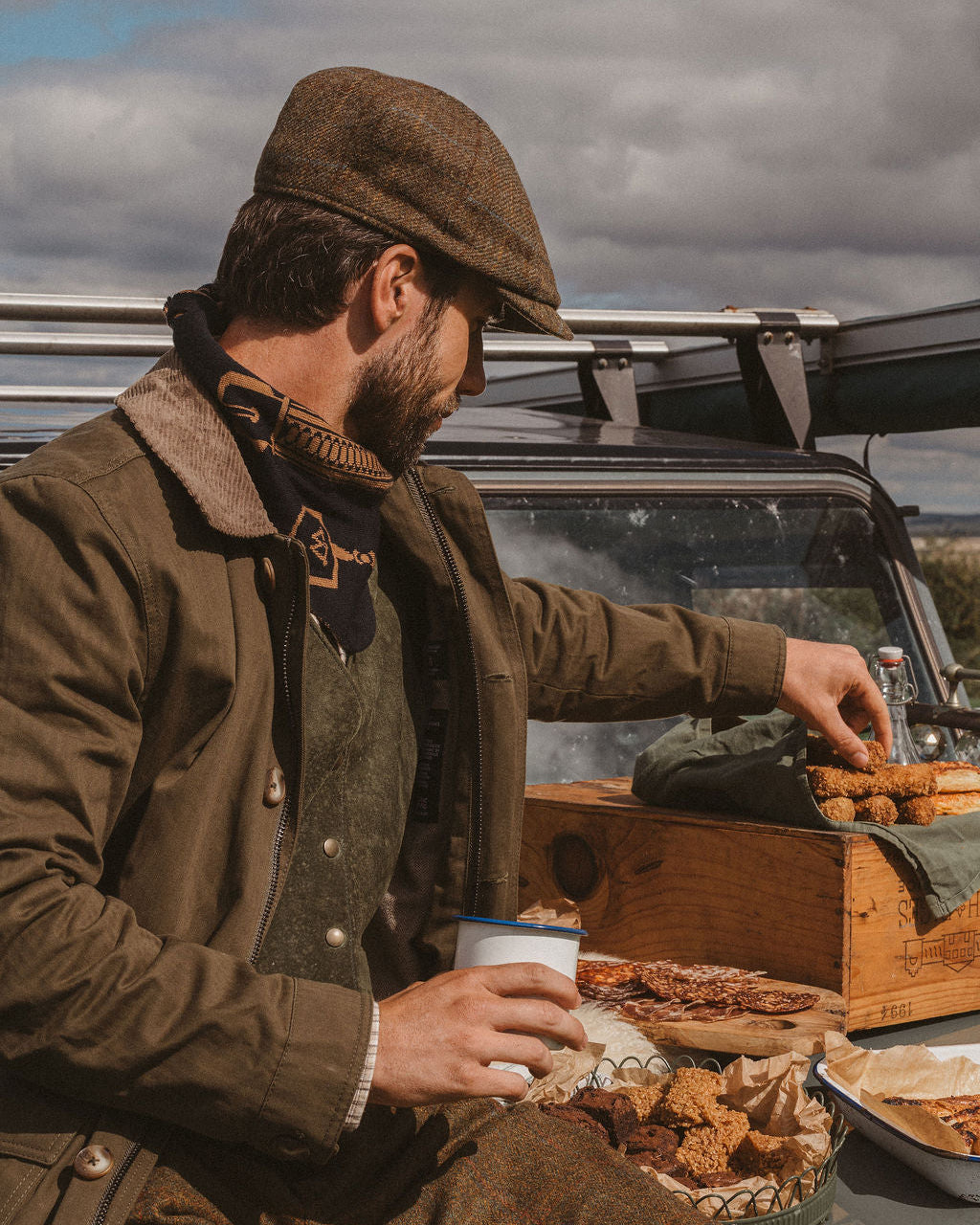 A man in rustic brown tweed clothing and a flat cap sits beside a vintage vehicle, reaching into a wooden food box outdoors under a cloudy sky.