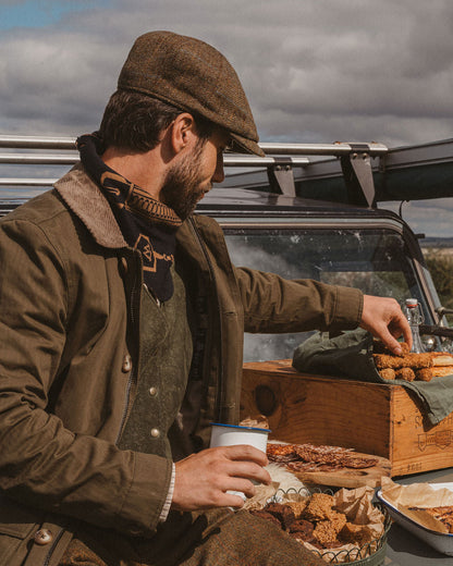 A man in rustic brown tweed clothing and a flat cap sits beside a vintage vehicle, reaching into a wooden food box outdoors under a cloudy sky.