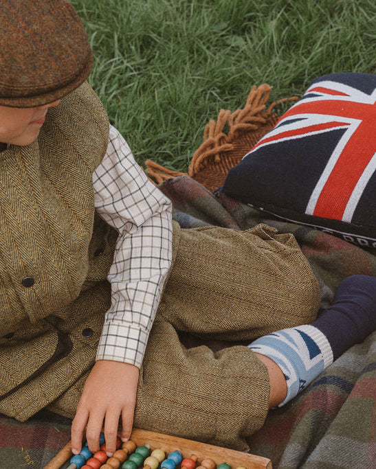 A child in vintage tweed clothing sits on a picnic blanket on grass, playing with colorful wooden beads beside a Union Jack pillow, wicker basket, and a plate of food.