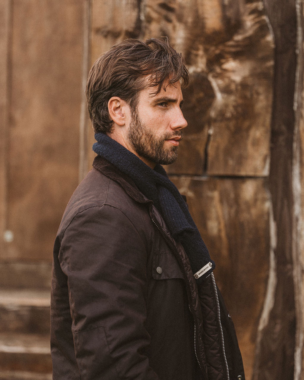 A man in profile wearing a dark brown jacket and a dark navy knit scarf, standing against a rustic wooden wall.