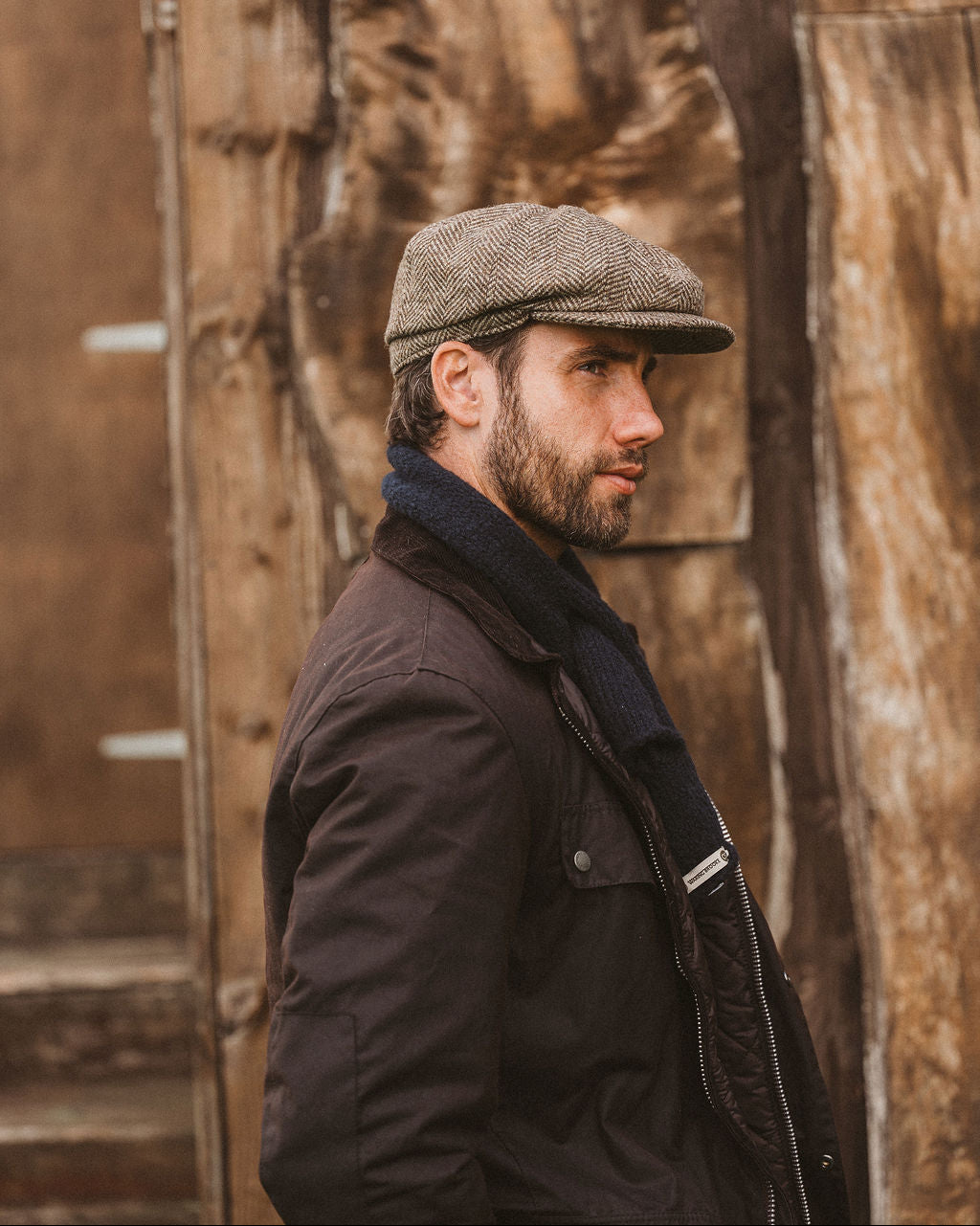 A man in profile wearing a brown tweed flat cap, a dark brown jacket, and a navy scarf, standing in front of a rustic wooden wall.