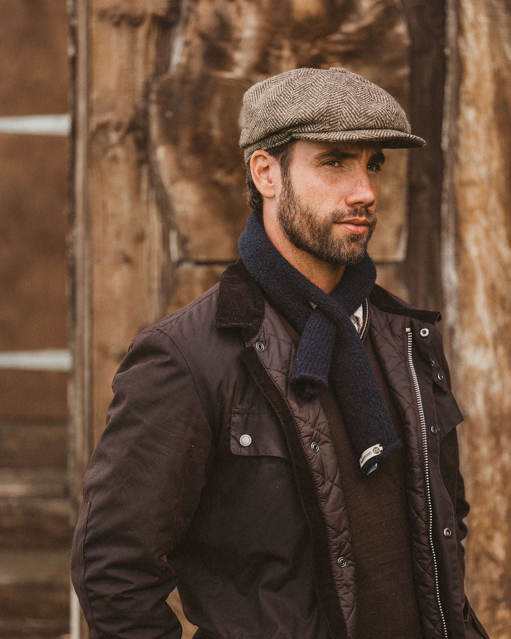 A man wearing a brown tweed flat cap, a dark brown waxed jacket, a navy blue knit scarf, and a brown quilted sweater, standing in front of a rustic wooden wall.