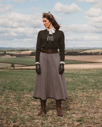 A woman stands outdoors in a grassy field under a blue sky with clouds, wearing a dark long-sleeve sweater over a white collared blouse, a long brown plaid skirt, black gloves, and brown boots.