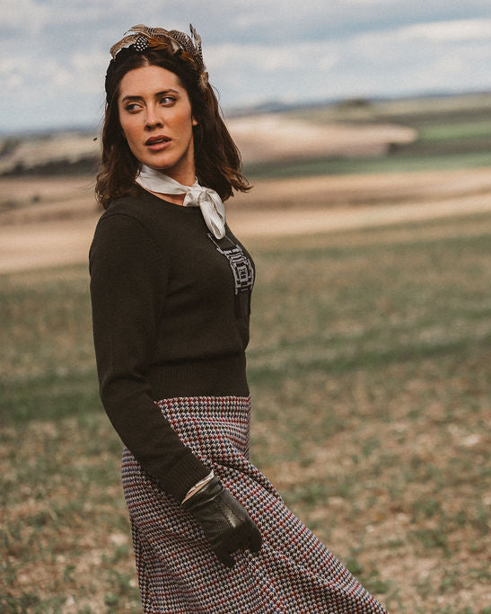 A woman stands in an open field wearing a dark long-sleeve top, a patterned skirt, a white neck scarf, and aviator-style sunglasses on her head.