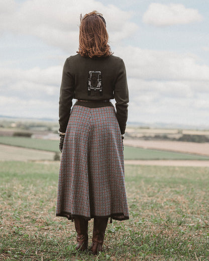 A woman viewed from behind stands in a grassy field under a cloudy sky, wearing a dark olive green long-sleeve top with a graphic print on the back and a brown, red, and blue plaid midi skirt.