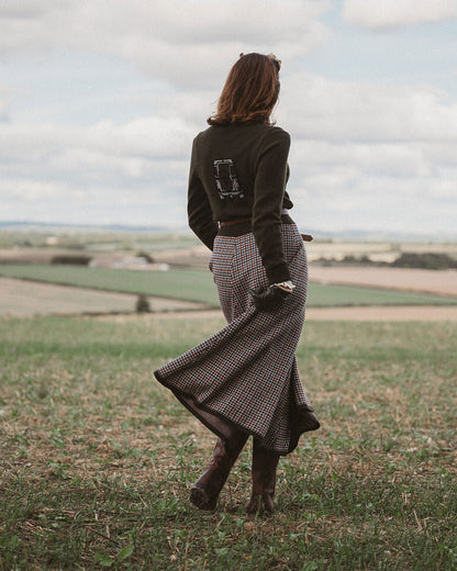 A woman seen from behind walking in an open grassy field under a cloudy sky, wearing a dark olive green long-sleeve top, a brown and beige plaid maxi skirt, and dark brown boots.