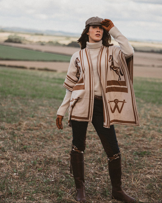 A woman stands in an open field wearing a cream knit poncho-style cardigan with brown embroidered motifs, a brown flat cap, dark pants, and tall brown boots.