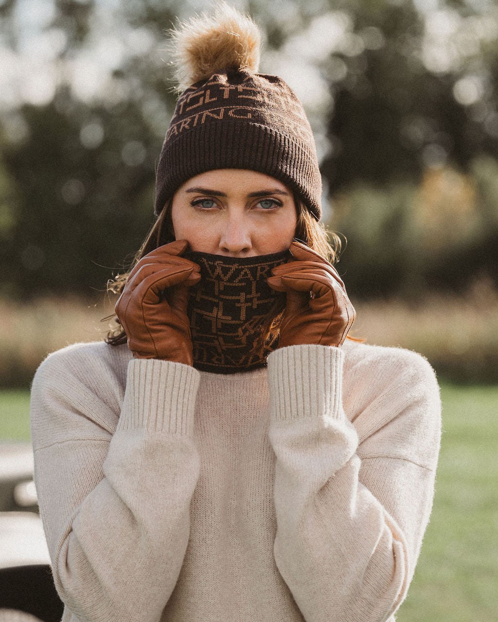 A woman outdoors wearing a dark brown knit beanie with a pom-pom, brown leather gloves, and a matching patterned neck gaiter over a cream knit sweater.