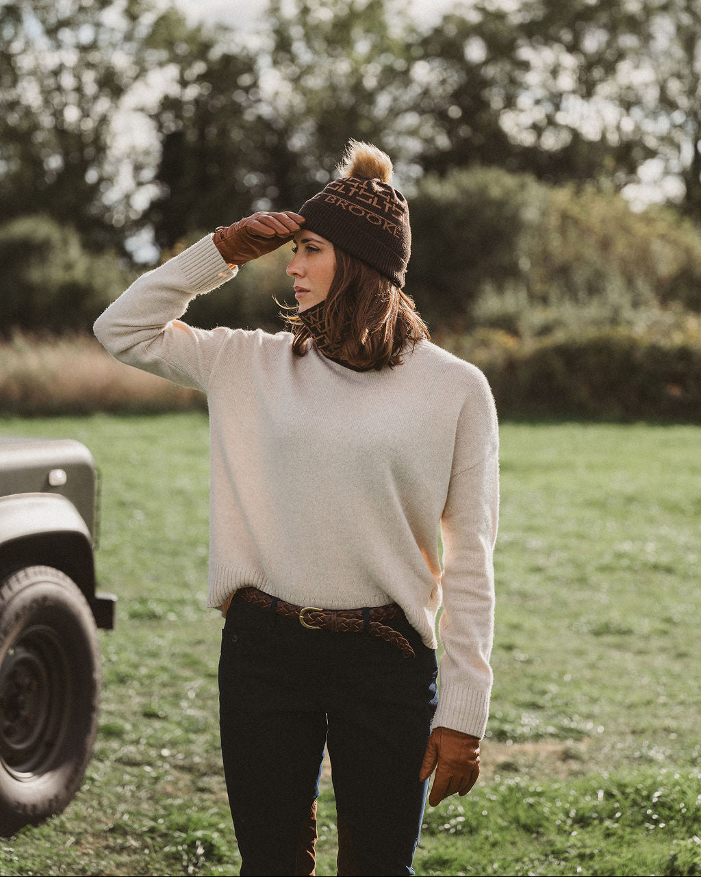 A woman standing outdoors in a grassy field wearing a cream knit sweater, a dark brown beanie with a pom-pom, black pants, and brown gloves, with trees and a vehicle blurred in the background.