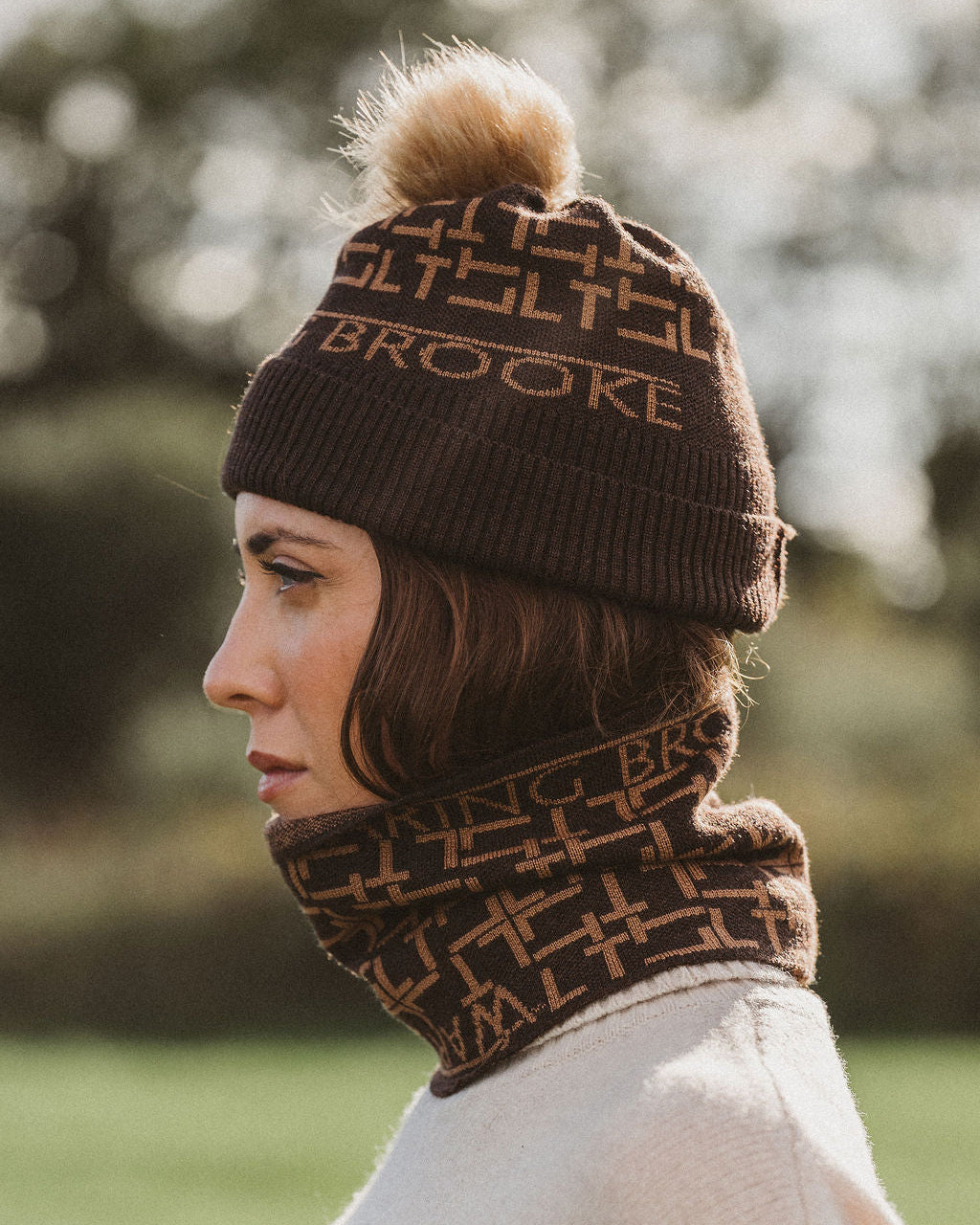 A woman in profile wearing a brown knit beanie with a matching pom-pom and a matching brown neck gaiter. The knit pattern features a repeating geometric design with the brand name 'BROOKE' clearly visible on the beanie.