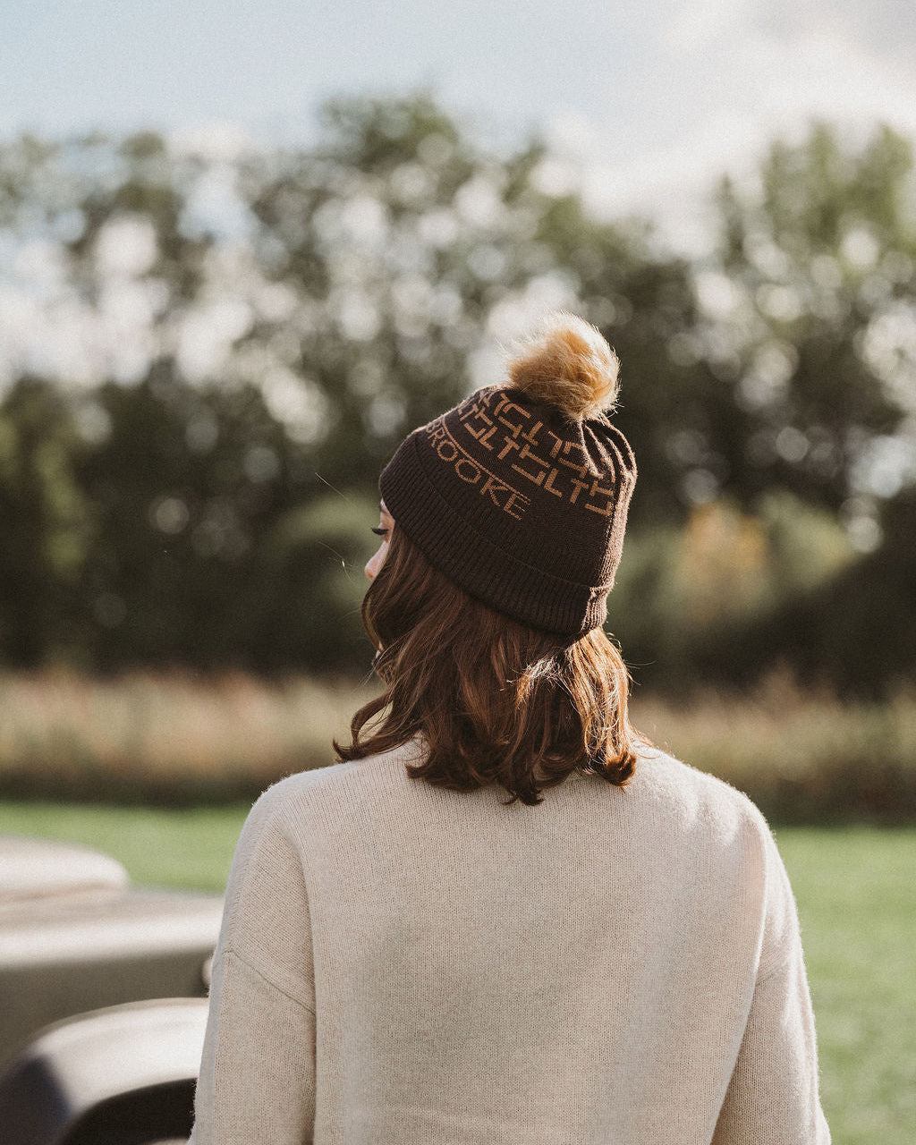 A woman viewed from behind wearing a dark brown knit beanie with a tan pom-pom and embroidered text, standing outdoors in a grassy area with trees in the background.