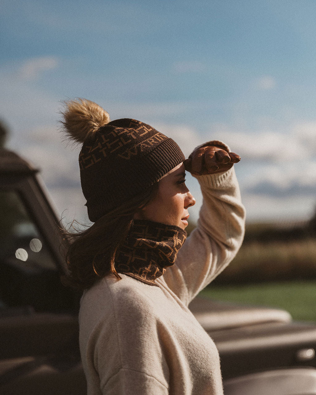A woman in a cream knit sweater, brown patterned beanie with a pom-pom, and a brown patterned scarf stands outdoors in profile against a blue sky, with a vintage car blurred in the background.